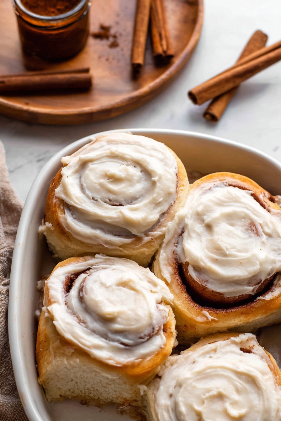 A cinnamon roll sits on a white plate with patterned edges, featuring multiple spiral layers of soft, golden brown dough wrapped around a dark cinnamon filling. The top layer is thickly coated with creamy white icing, slightly melting and dripping down the sides. The bottom shows some syrupy cinnamon mixture oozing out, adding a sticky texture. In the background, there is a wooden bowl with several cinnamon sticks inside, all set on a white marbled surface. photo taken with an iphone --ar 2:3 --v 7