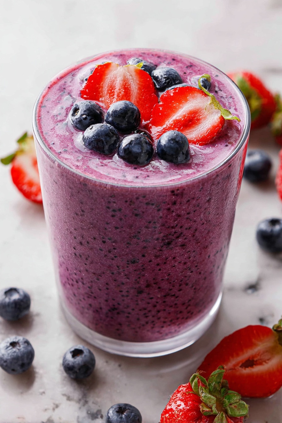 A clear glass filled with a thick purple smoothie that has a textured surface showing tiny berry seeds. The top layer of the smoothie is garnished with whole blueberries and two halves of bright red strawberries with green caps, lying flat on the smoothie surface. The glass is placed on a white marbled surface with scattered blueberries and strawberry halves around it. The lighting is bright, highlighting the fresh look and colors of the fruit. photo taken with an iphone --ar 2:3 --v 7