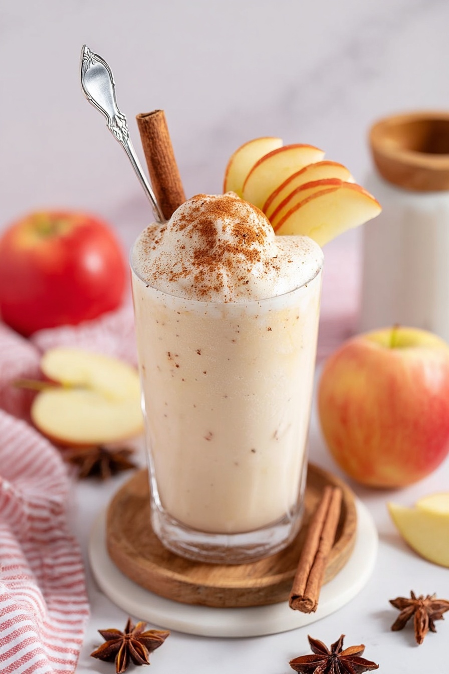 A tall clear glass filled with a creamy white drink that has a smooth, slightly frothy texture with light brown specks inside, placed on a round wooden coaster. On top of the drink, three thin yellow apple slices lean against a cinnamon stick and star anise, all held together by a silver cocktail pick. Surrounding the glass are whole red apples, star anise scattered, a cinnamon stick, and a white bowl with star anise on a white marbled surface. A white jar with a wooden lid and a red and white striped cloth are in the blurred background. photo taken with an iphone --ar 2:3 --v 7