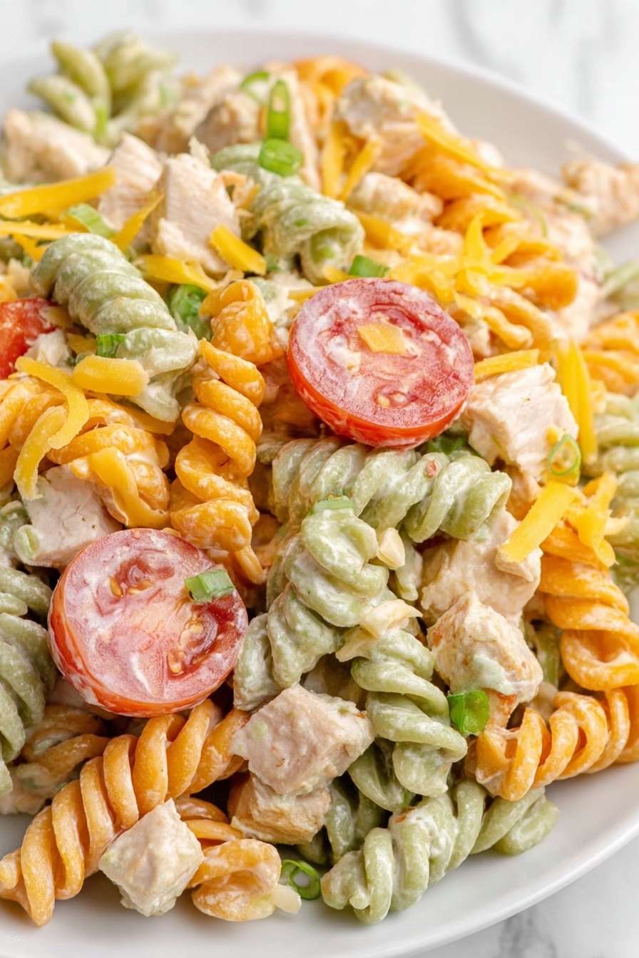The dish shows a close-up of tri-color rotini pasta salad with green, orange, and white twisted pasta pieces mixed with creamy dressing. There are sliced red tomatoes with a smooth texture placed among the pasta, topped with small cubes of light beige chicken and bits of green onions scattered throughout. Shredded yellow cheddar cheese is mixed in, adding extra color and texture. The salad is served on a white plate, with a white marbled surface in the background. photo taken with an iphone --ar 2:3 --v 7