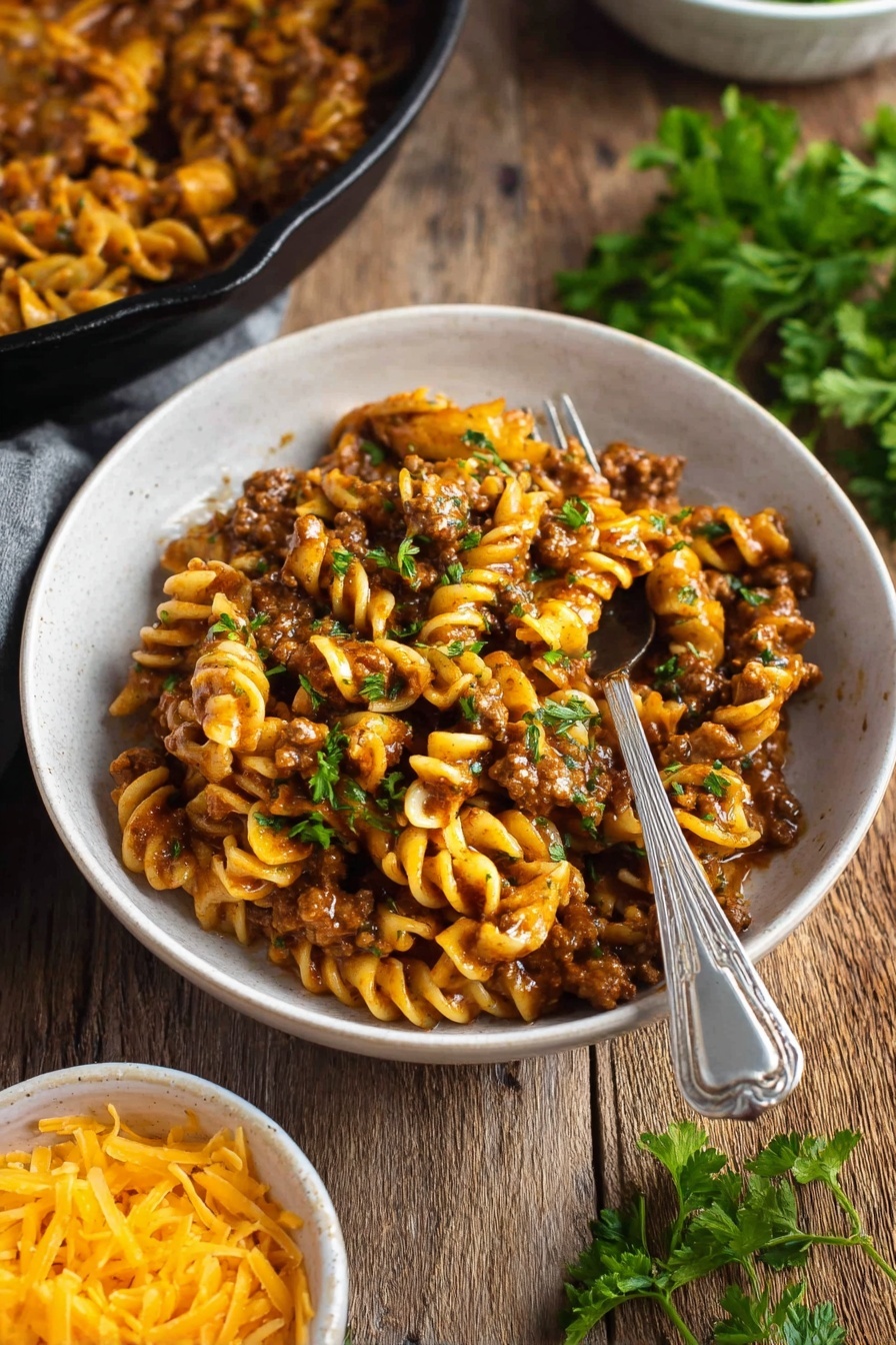 Vegan Cheeseburger Pasta - A black cast iron pan holds a creamy pasta dish with three layers: the bottom has a sauce mixed with ground plant-based meat that is dark brown and slightly chunky, the middle layer features short, spiral pasta in a light yellow-orange color coated in sauce, and the top layer is sprinkling of small green parsley pieces for garnish. The pan is placed on a black and white striped cloth on a wooden surface with uncooked spiral pasta scattered around. To the left of the pan, a white bowl contains shredded yellow cheddar cheese, and above is a package of plant-based meat. The background is a white marbled texture. Photo taken with an iphone --ar 2:3 --v 7