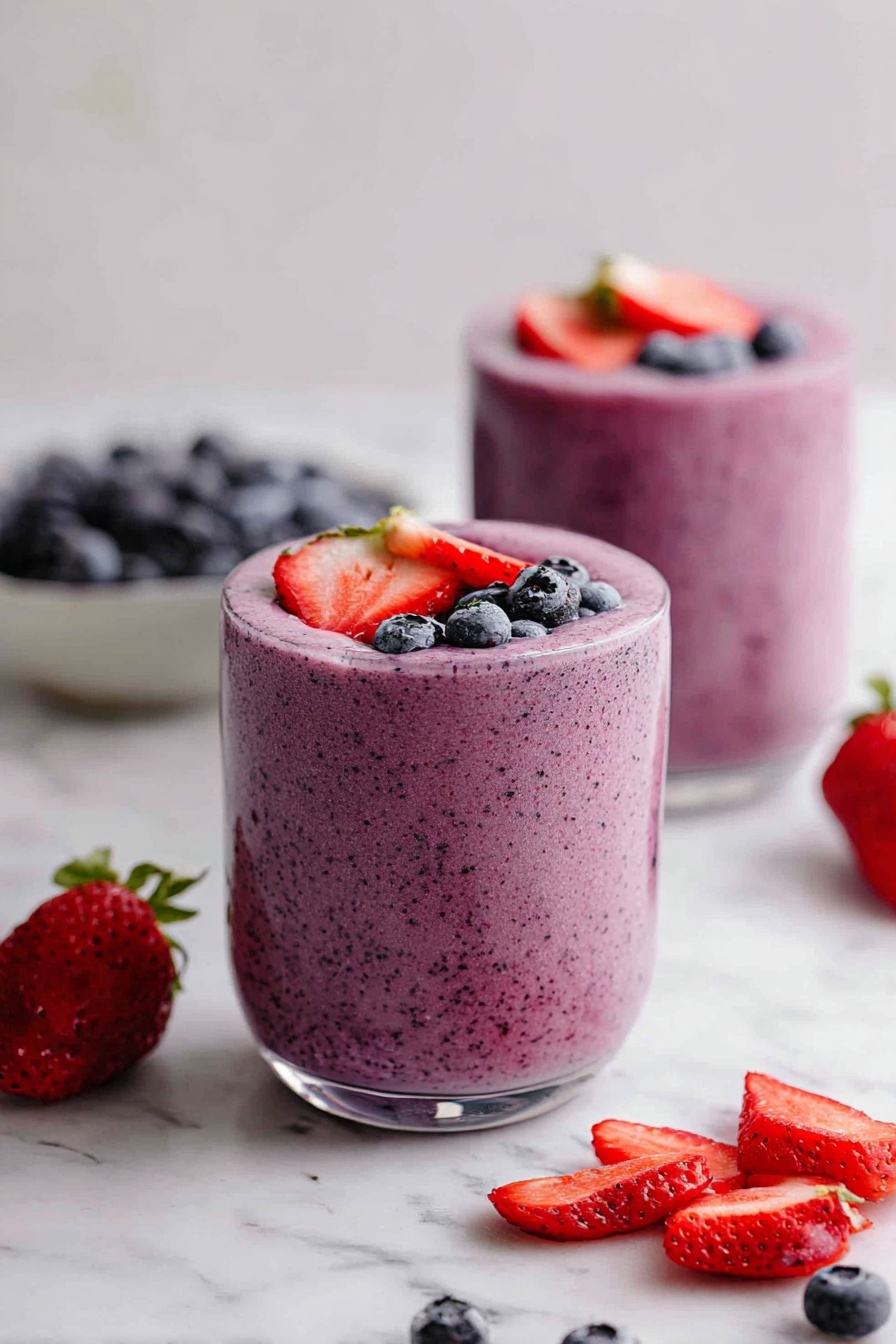 Two clear round glass cups filled with a thick pink smoothie made from blended berries. Each cup is topped with whole blueberries and halved strawberries with green tops. Around the glasses, there are extra fresh strawberries and blueberries, some placed inside two white bowls and some scattered on a white marbled surface. A piece of white lace fabric is partly visible near the top right corner. The scene is bright and fresh, with the colors of red strawberries and dark blue blueberries standing out against the pink smoothie and white marble background. photo taken with an iphone --ar 2:3 --v 7