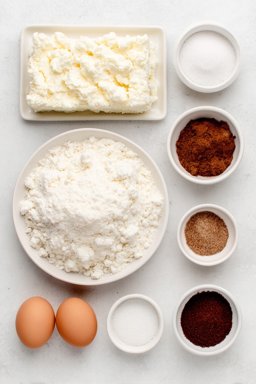 Flat lay of a block of cold cream cheese next to a small white bowl filled with granulated white sugar, a small white bowl with ground ginger, a small white bowl with ground cinnamon, a small white bowl with ground allspice, a small white bowl with ground nutmeg, a small white bowl with ground cloves, a mound of all-purpose flour on a white ceramic plate, a stick of softened unsalted butter, a small pile of packed light brown sugar, two whole uncracked brown eggs, a small white bowl with dark unsulphured molasses, and a small white bowl with vanilla extract placed on a clean white marble surface, soft natural light, photo taken with an iPhone, professional food photography style, fresh ingredients, white ceramic bowls, no bottles, no duplicates, no utensils, no packaging --ar 2:3 --v 7 --p m7354615311229779997