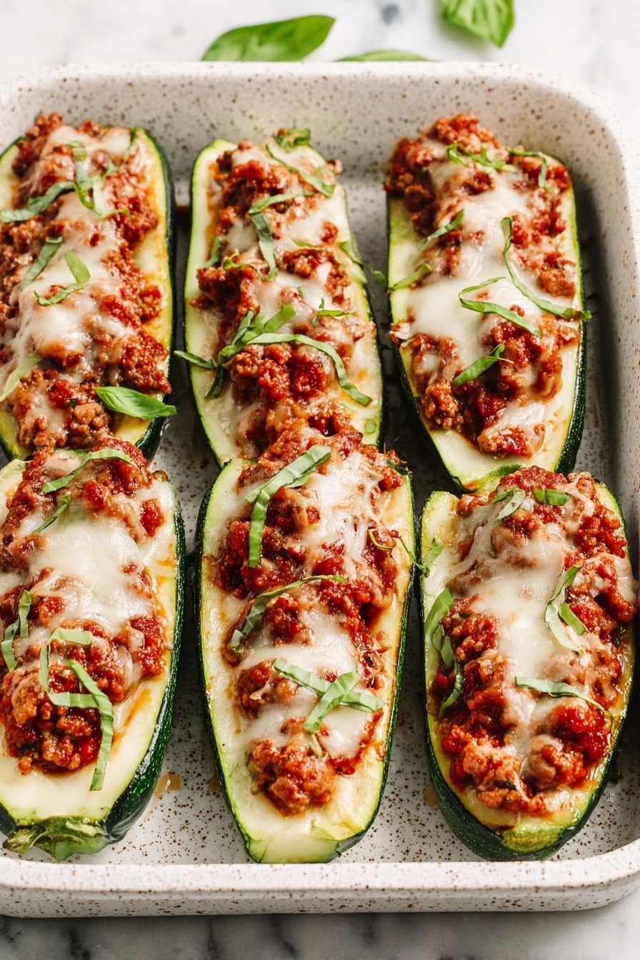 The image shows a white speckled baking dish with six zucchini boats arranged in two rows. Each zucchini boat is hollowed out and filled with a mixture of reddish-brown cooked ground meat and tomato sauce. On top of the filling is a layer of melted white cheese that looks smooth and slightly browned in places. Garnished sprinkle of chopped fresh green basil leaves is scattered across the boats. The baking dish sits on a white marbled surface, with parts of other dishes and fresh herbs faintly visible around the edges. Photo taken with an iphone --ar 2:3 --v 7