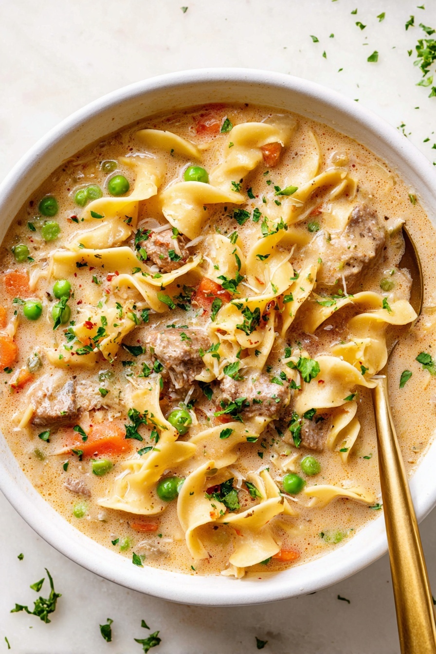 The image shows a close-up of a white bowl filled with creamy beef noodle soup. The first layer is a smooth, light beige creamy broth. Floating in the broth are wide yellow egg noodles with a slight curl and soft texture. There are medium-sized brown beef chunks spread throughout the noodles. Bright green peas and orange carrot pieces add pops of color. Small bits of chopped green herbs are sprinkled on the top layer to add freshness. A silver spoon is partially visible dipped into the soup from the left side. The bowl is placed on a white marbled surface. photo taken with an iphone --ar 2:3 --v 7