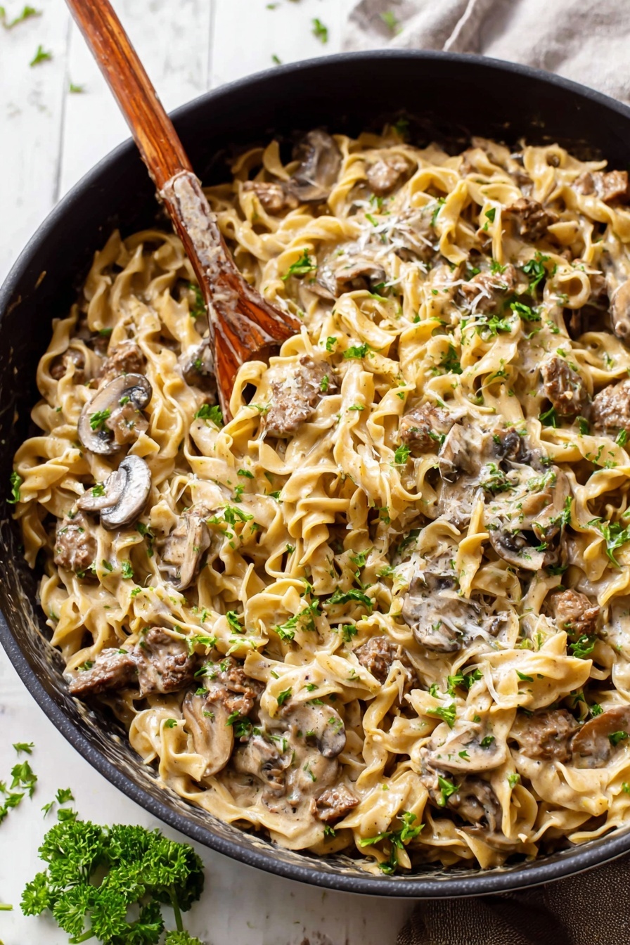 A large black pan filled with creamy noodles mixed with slices of brown mushrooms and small chunks of browned meat, all covered in a thick light beige sauce. The noodles are twisted and soft, scattered evenly throughout the pan. Small green parsley leaves are sprinkled over the top, adding bits of fresh color. A wooden-handled spoon is stuck into the noodles, ready for serving. The pan rests on a white marbled surface with some parsley pieces scattered around. photo taken with an iphone --ar 2:3 --v 7
