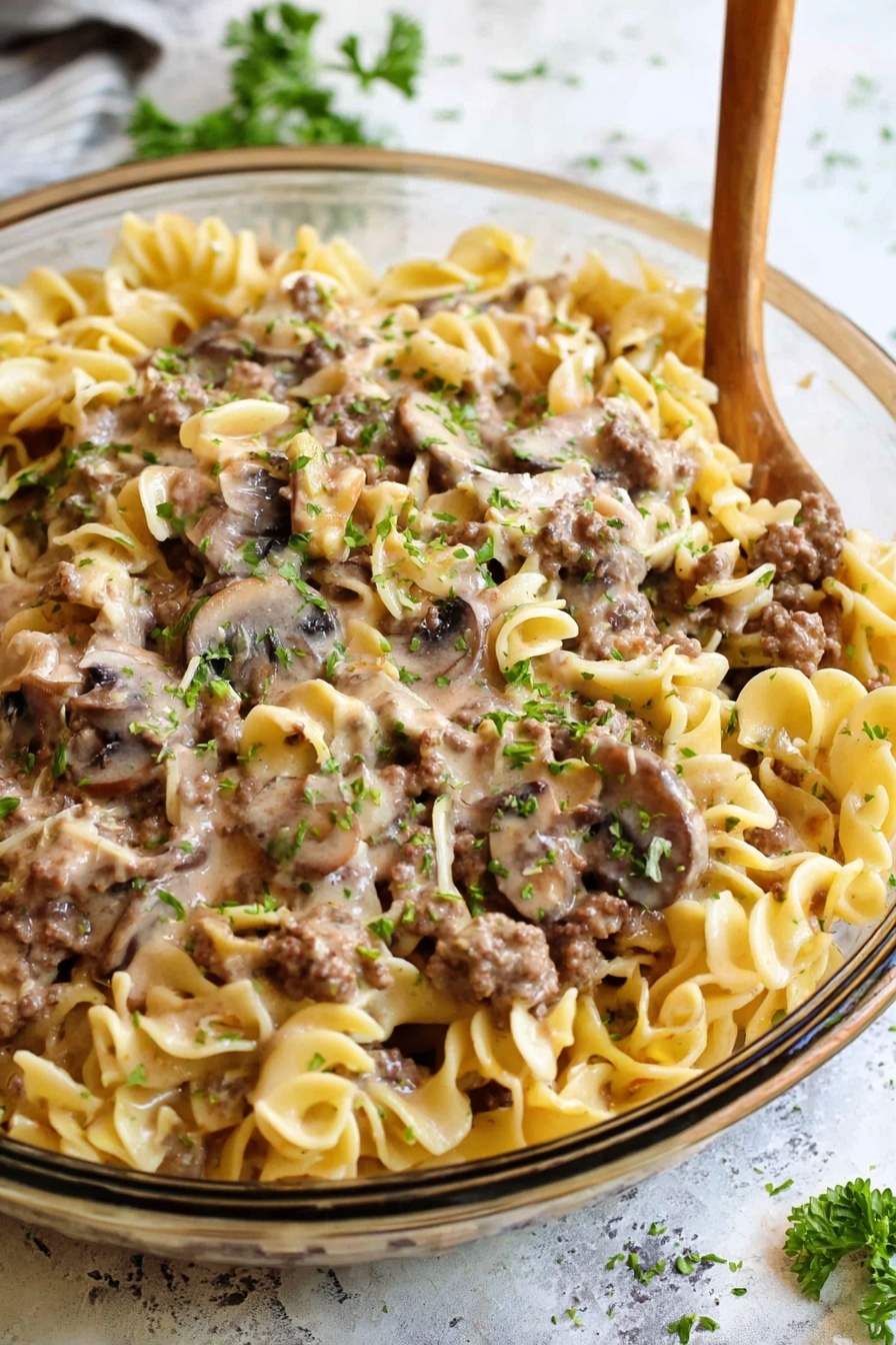 A large bowl filled with creamy pasta that has a base layer of wide, flat noodles in pale yellow. Mixed in are browned pieces of ground meat and cooked mushroom slices in dark brown with a shiny texture. The dish is coated in a light beige creamy sauce, sprinkled with small green parsley garnishes on top. A gold spoon is placed inside the bowl, with a woman's hand holding the bowl from below. The background and surface are white marbled texture with soft natural lighting. photo taken with an iphone --ar 2:3 --v 7