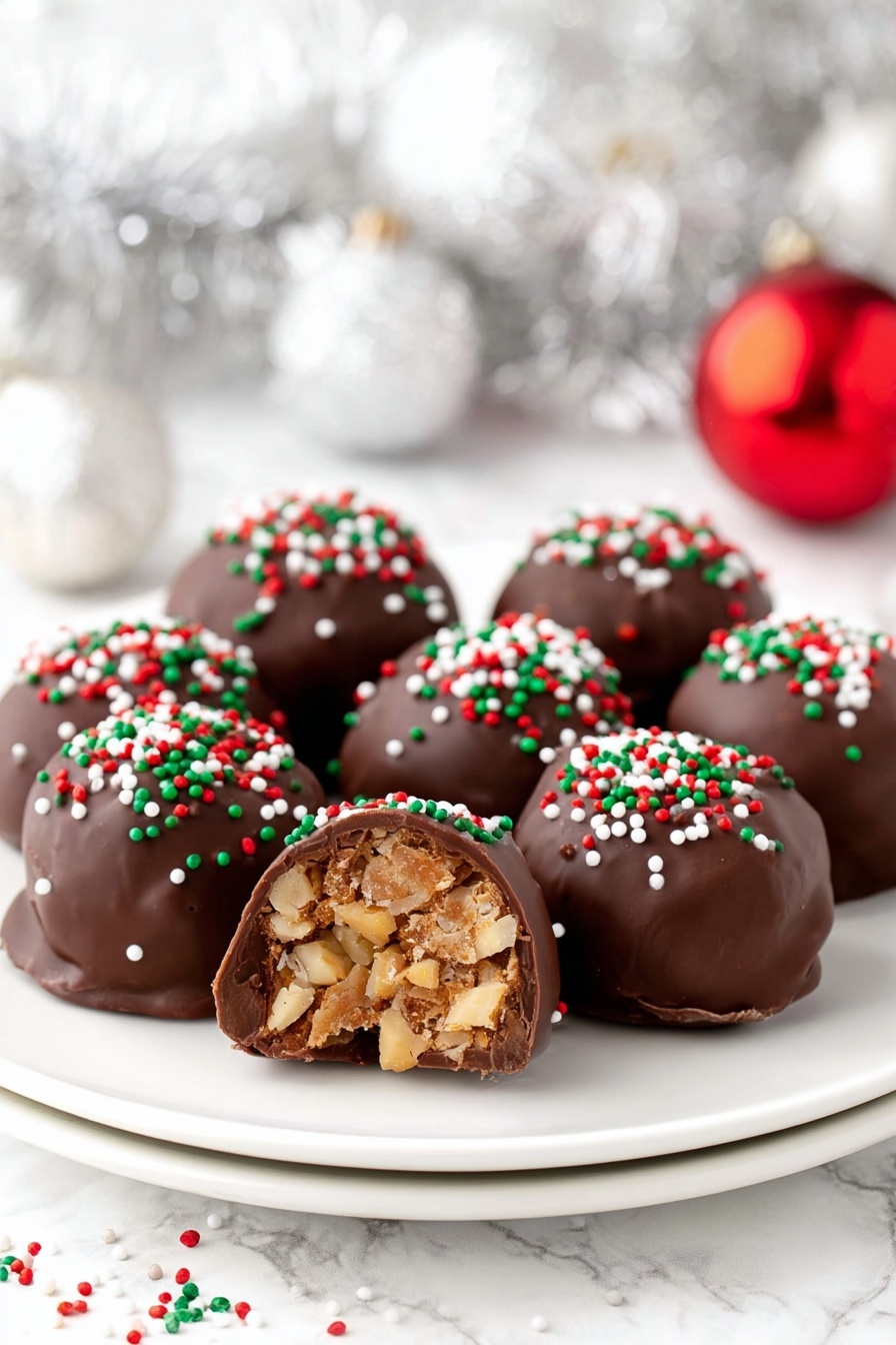A white plate holds eight round chocolate-covered balls, each topped with small red, green, and white sprinkles. One ball is cut in half and placed in front, showing a light brown inside with pieces of nuts. The chocolate coating is smooth and shiny, slightly melting at the bottom edges. The background has a white marbled texture with soft, out-of-focus silver decorations and a red ornament. Sprinkles are scattered lightly on the plate and surface. photo taken with an iphone --ar 2:3 --v 7