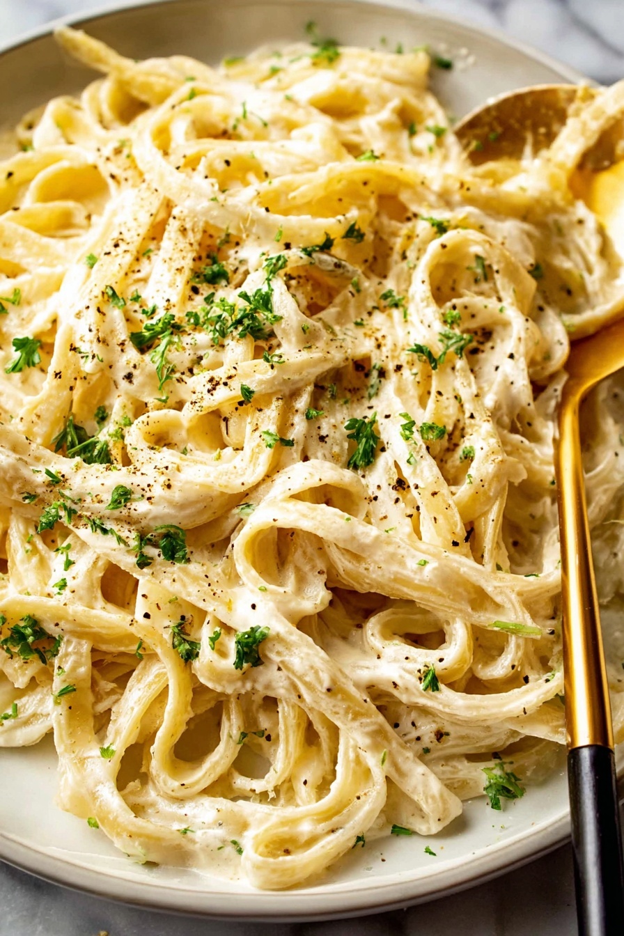 The image shows a close-up of a bowl filled with creamy pasta. The pasta appears to be fettuccine, thick and coated evenly with a smooth, light cream sauce. On top, finely chopped green herbs and a sprinkle of black pepper add a touch of color and texture. The bowl is white with small brown speckles, and a gold fork is stuck into the pasta on the left side. The background has a white marbled texture. photo taken with an iphone --ar 2:3 --v 7