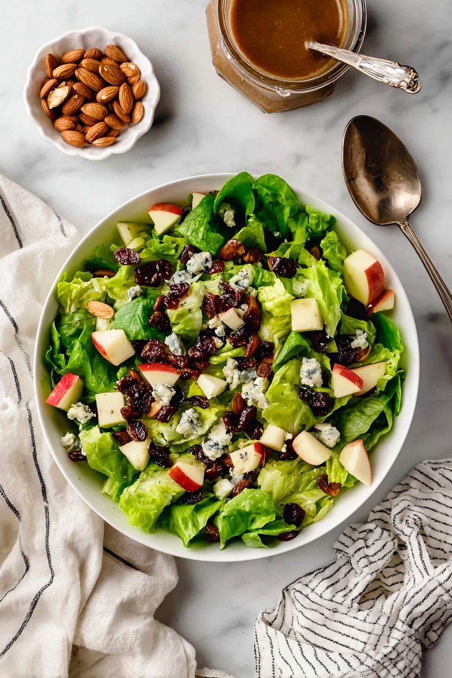 A fresh salad served in a white bowl placed on a white marbled surface shows bright green leafy lettuce as the first layer. On top, there are small cubes of red and yellow apples spread evenly, mixed with dark red dried cranberries. Whole roasted almonds add a crunchy texture and a warm brown color, scattered among the other ingredients. Small bits of blue cheese with its white and dark blue veins sit atop the salad, giving a creamy look. The mix of colors and textures makes the salad look fresh and tasty, with a silver spoon visible near the top right corner. Photo taken with an iphone --ar 2:3 --v 7
