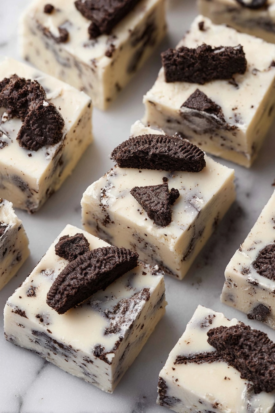 The image shows several square pieces of white fudge with dark chocolate cookie chunks mixed in and placed on top. Each piece has a creamy white base layer with uneven dark cookie bits embedded throughout. The top is decorated with larger cookie pieces that have a rough texture, creating a contrast against the smooth fudge. The fudge squares are arranged closely together on a white marbled surface, with a soft light highlighting the creamy texture and the cookie details. photo taken with an iphone --ar 2:3 --v 7