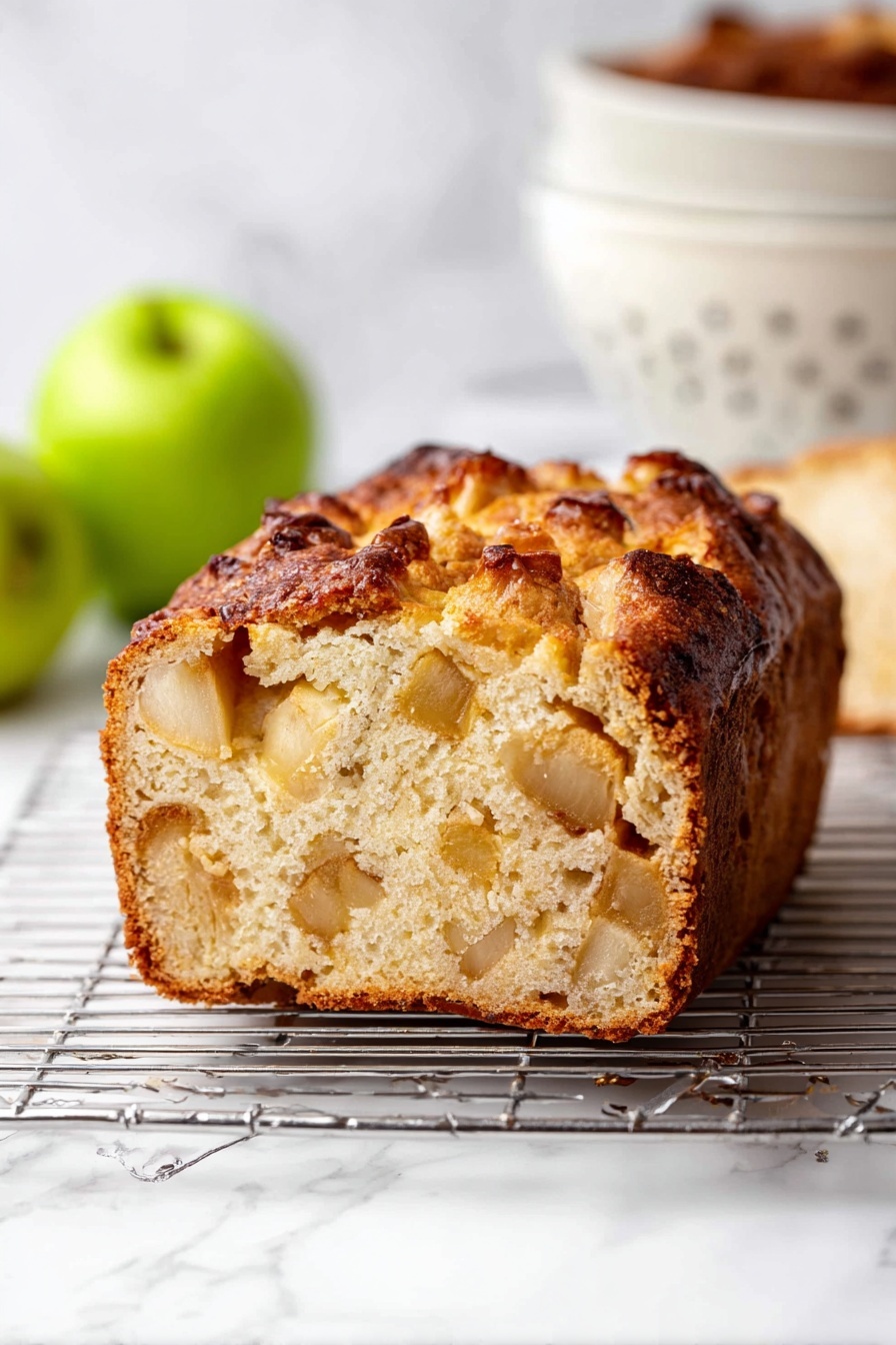 The image shows a thick slice of golden brown bread with a soft and fluffy inside. The bread has visible chunks of light tan apple pieces spread throughout its two main layers, which show a dense yet airy texture. The top layer has a darker caramelized crust with a slightly rough and uneven surface. The bread sits on a metal cooling rack placed on a white marbled surface. In the background, there is a white colander and two green apples, adding fresh colors to the scene. Photo taken with an iphone --ar 2:3 --v 7