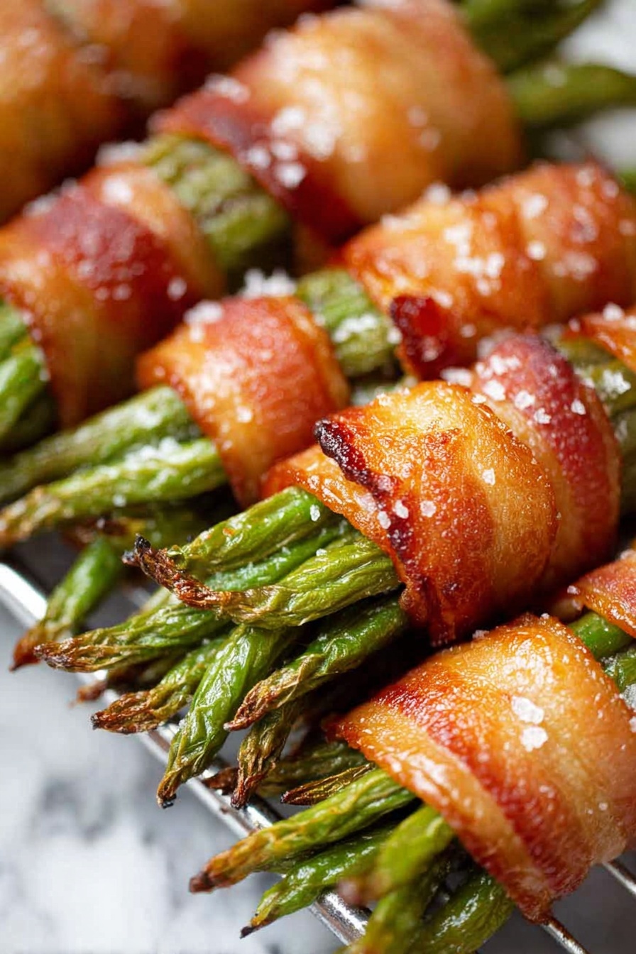 The image shows five bundles of green beans wrapped tightly with crispy, golden-brown bacon strips. Each bundle has about 7-10 green beans, with their slightly charred tips sticking out from both ends of the bacon wrap. The bacon looks shiny and textured with visible salt flakes on top. These bundles rest on a metal cooling rack inside a baking tray lined with a browned parchment paper that has dark spots from cooking. The edge of the tray has a crisp folded rim and a navy, white striped cloth is partly visible at the bottom left corner. The whole scene is set against a white marbled texture photo taken with an iphone --ar 2:3 --v 7