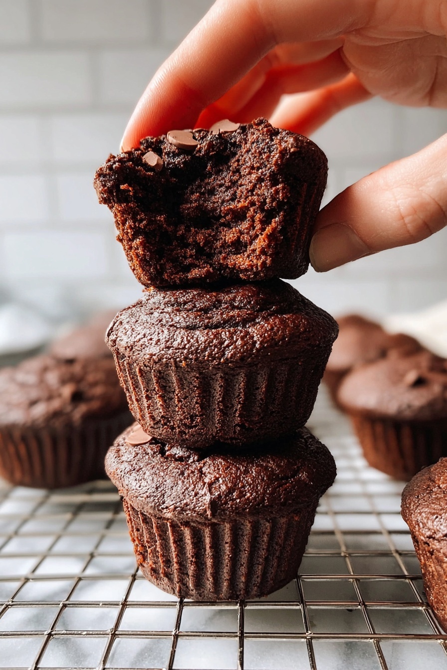 The image shows seven dark brown chocolate muffins with a rough, cracked top texture. Six muffins are placed on a metal cooling rack, with one muffin lying on its side showing a ridged wrapper and soft inside. The muffins look dense and moist with some visible chunks of chocolate on top. The cooling rack sits on a white marbled surface scattered with small chocolate pieces and pale yellow flower petals. In the bottom left corner, there is a white bowl filled with dark chocolate chips. The lighting is natural, highlighting the rich color and texture of the muffins. photo taken with an iphone --ar 2:3 --v 7