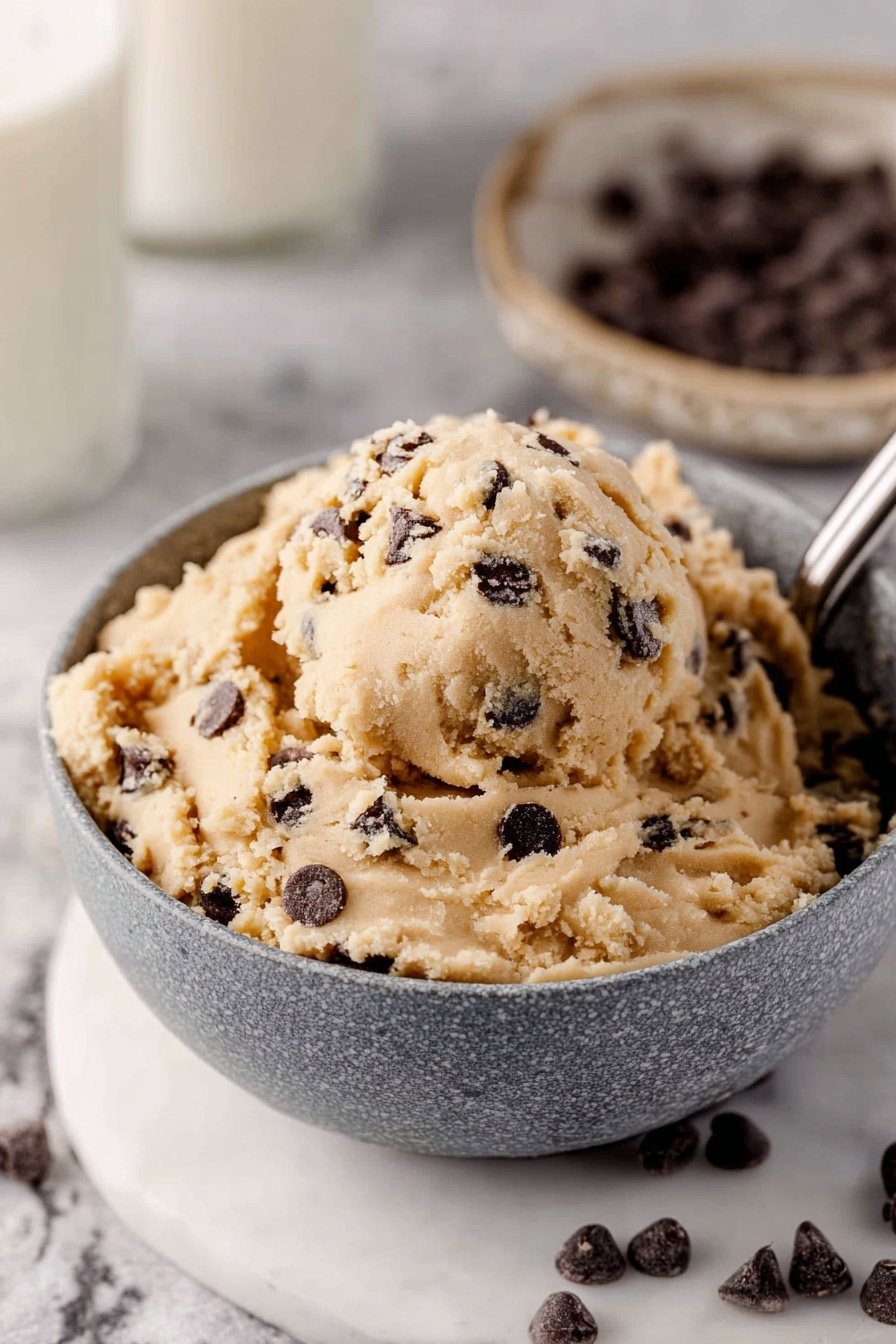 A close-up view of a grey bowl filled with creamy, pale tan cookie dough that has many small dark brown chocolate chips mixed in. The dough looks soft and a bit crumbly, with a round scoop of it resting on a metal spoon sticking out from the right side. The bowl sits on a white marbled surface, and in the background, there is a glass of milk that is partly out of focus along with a beige speckled plate that holds more chocolate chips. Some chocolate chips are scattered around the bowl on the marbled surface. photo taken with an iphone --ar 2:3 --v 7