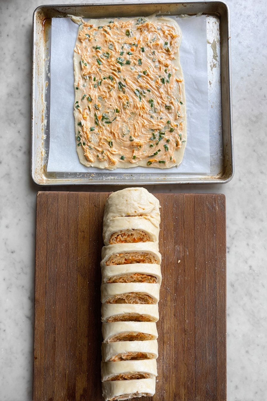 There is a white plate with a beaded edge holding nine golden-brown pastry pinwheels, each topped with small green onion pieces. The pinwheels have visible layers of flaky dough with an orange filling spread inside, showing a slightly rough texture. A clear glass bowl of white dipping sauce sits in the middle of the plate. A woman's hand is holding one of the pinwheels, dipping it partly into the sauce. The plate is placed on a white marbled surface with a striped cloth nearby. photo taken with an iphone --ar 2:3 --v 7