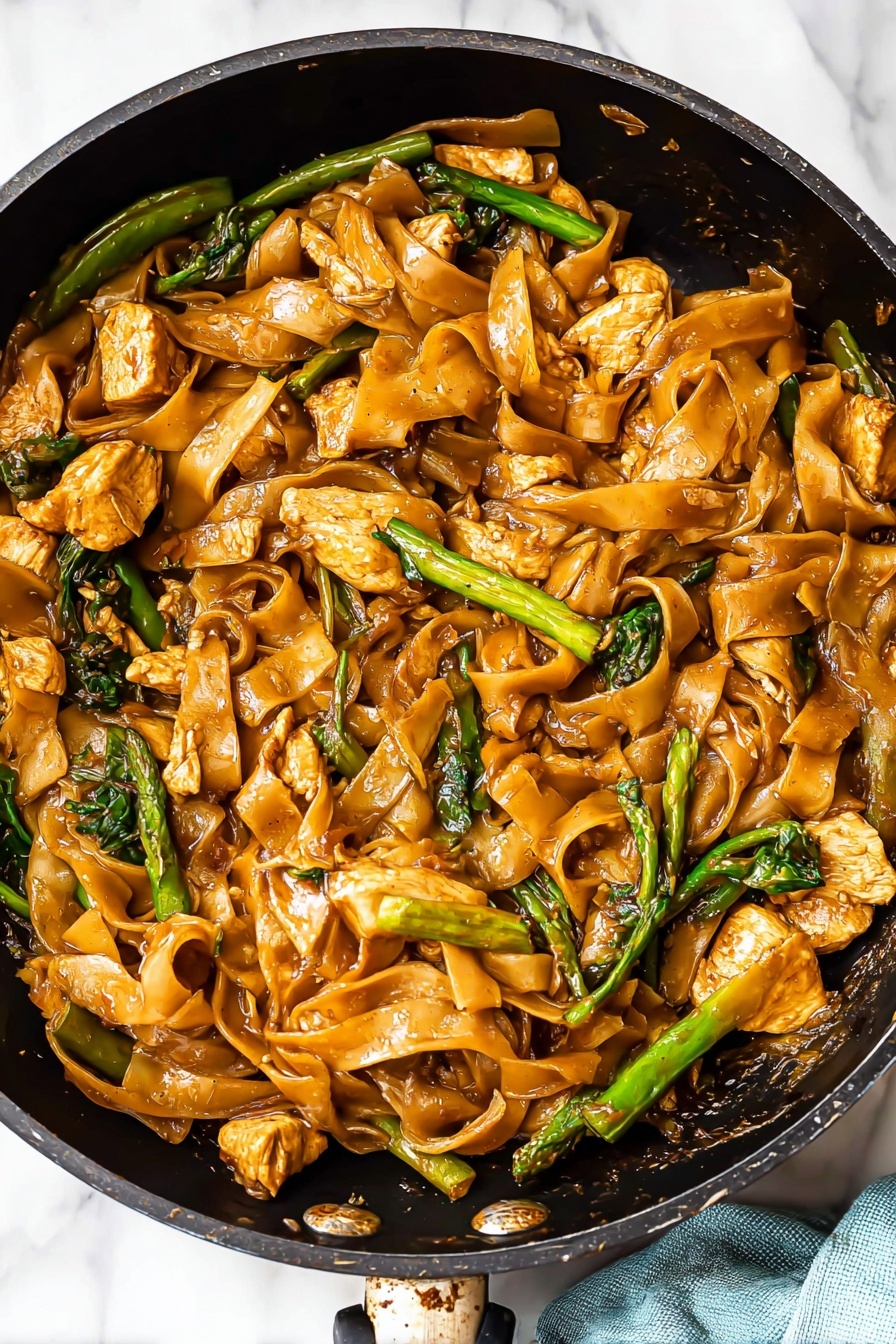 The image shows a pan full of flat, wide noodles cooked in a shiny brown sauce that covers them evenly, giving a slightly glossy texture. Mixed in are chunky pieces of light brown cooked chicken and bright green vegetables like leafy greens and long slices of green beans. The noodles have some crinkled edges and vary slightly in shade due to the sauce. The pan is black inside and sits on a white marbled surface, with a glimpse of a light blue cloth nearby. photo taken with an iphone --ar 2:3 --v 7