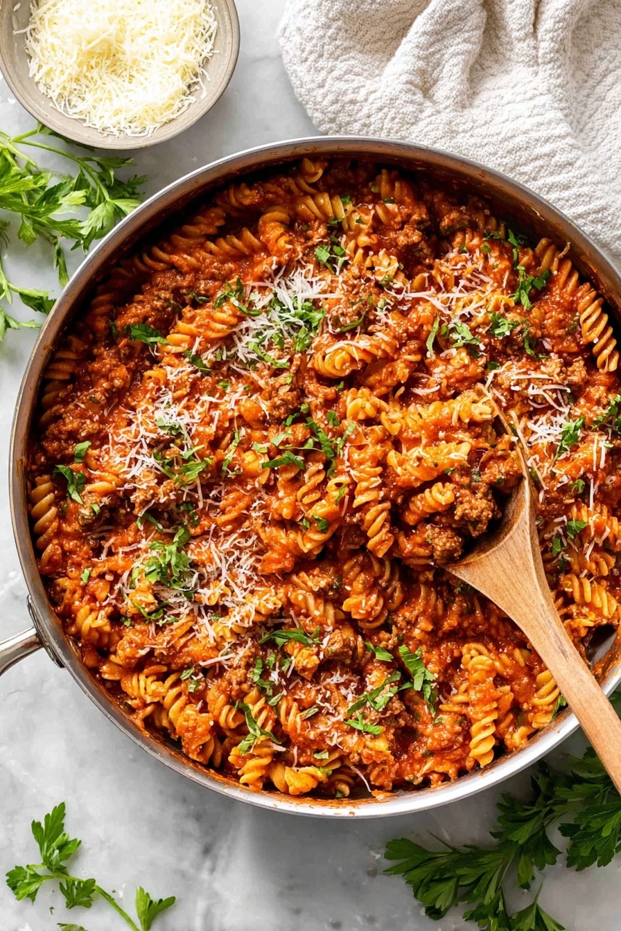 A large silver pan filled with a thick mixed dish of spiraled pasta and chunky red tomato sauce with browned ground meat, topped with small green parsley leaves and sprinkled with fine white cheese shreds. A wooden spoon inside the pan lifts a scoop of the pasta mixture. The pan is on a white marbled surface with some green parsley sprigs scattered nearby and a small bowl with shredded white cheese in the upper left. A soft white cloth lies folded in the upper right corner. photo taken with an iphone --ar 2:3 --v 7