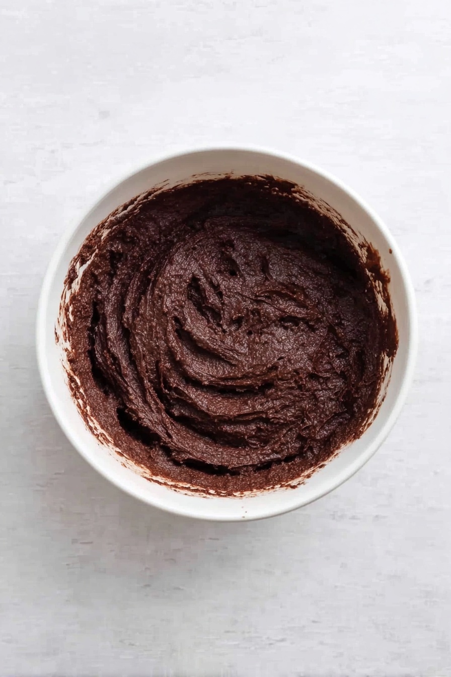 The image shows two stacked square brownies with three layers each: a thick dark brown chocolate layer at the bottom and middle, and a creamy white cheesecake layer on top and between the chocolate layers. Swirls of deep red raspberry sauce are mixed into the cheesecake layers on top, creating a marbled pattern. In the background on a white marbled surface, there are more brownies and a small white bowl filled with fresh red raspberries. The focus is on the front stacked brownies showing the rich texture of the chocolate and smooth swirled cheesecake. photo taken with an iphone --ar 2:3 --v 7