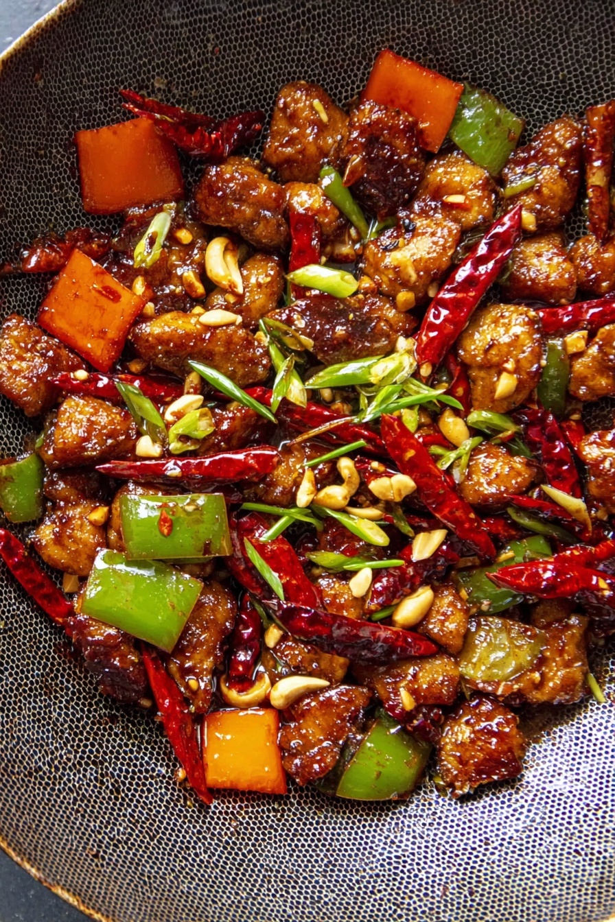 The image shows a close-up view of a pan filled with stir-fried pieces of brown-colored meat coated in a shiny sauce, mixed with bright red dried chili peppers scattered throughout. Chopped green and orange bell peppers add vibrant colors and a slightly crisp texture, while small pieces of chopped green onions and pale peanuts are sprinkled on top as garnishes. The pan has a dark, textured surface with a honeycomb pattern, and the dish looks hot and freshly cooked. Photo taken with an iphone --ar 2:3 --v 7