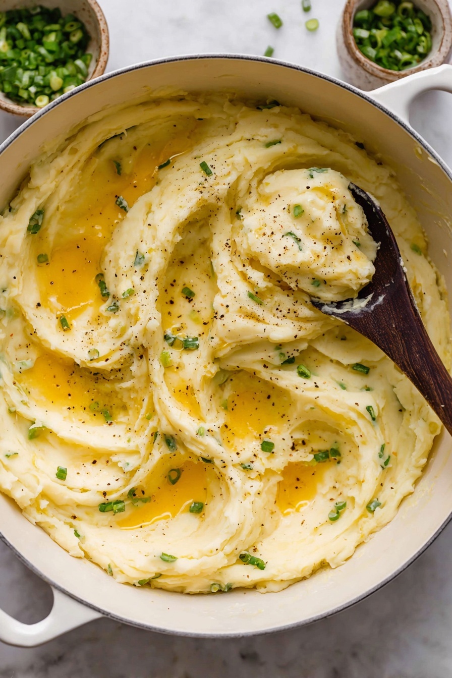 A large white pot filled with creamy mashed potatoes that are smooth and slightly fluffy in texture, showing soft swirls and peaks. The mashed potatoes have small green pieces of chopped scallions mixed in, adding flecks of color. There are pools of melted golden butter spread unevenly on the surface, glistening and blending into the potatoes. Freshly ground black pepper is sprinkled lightly across the top. A dark wooden spoon is scooping up a portion from the center, showing the thick, soft texture of the mash. The pot is set on a white marbled surface with small bowls of green onions and sauce nearby. photo taken with an iphone --ar 2:3 --v 7