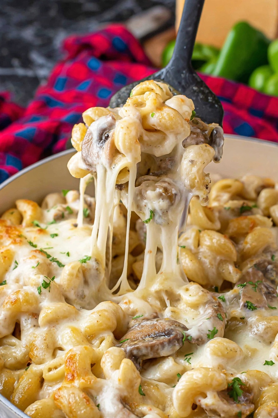 The image shows a close-up of a white pan filled with pasta in a creamy, melted cheese sauce. The pasta is light beige with smooth and slightly curled edges, covered in a thick layer of melted cheese that stretches as it is lifted with a spoon. There are pieces of brown mushrooms mixed in, adding texture to the creamy cheese and pasta. Small green parsley bits are scattered on top, adding color contrast. The background has a red and white checkered cloth and some blurred green vegetables on a white marbled surface. Photo taken with an iphone --ar 2:3 --v 7